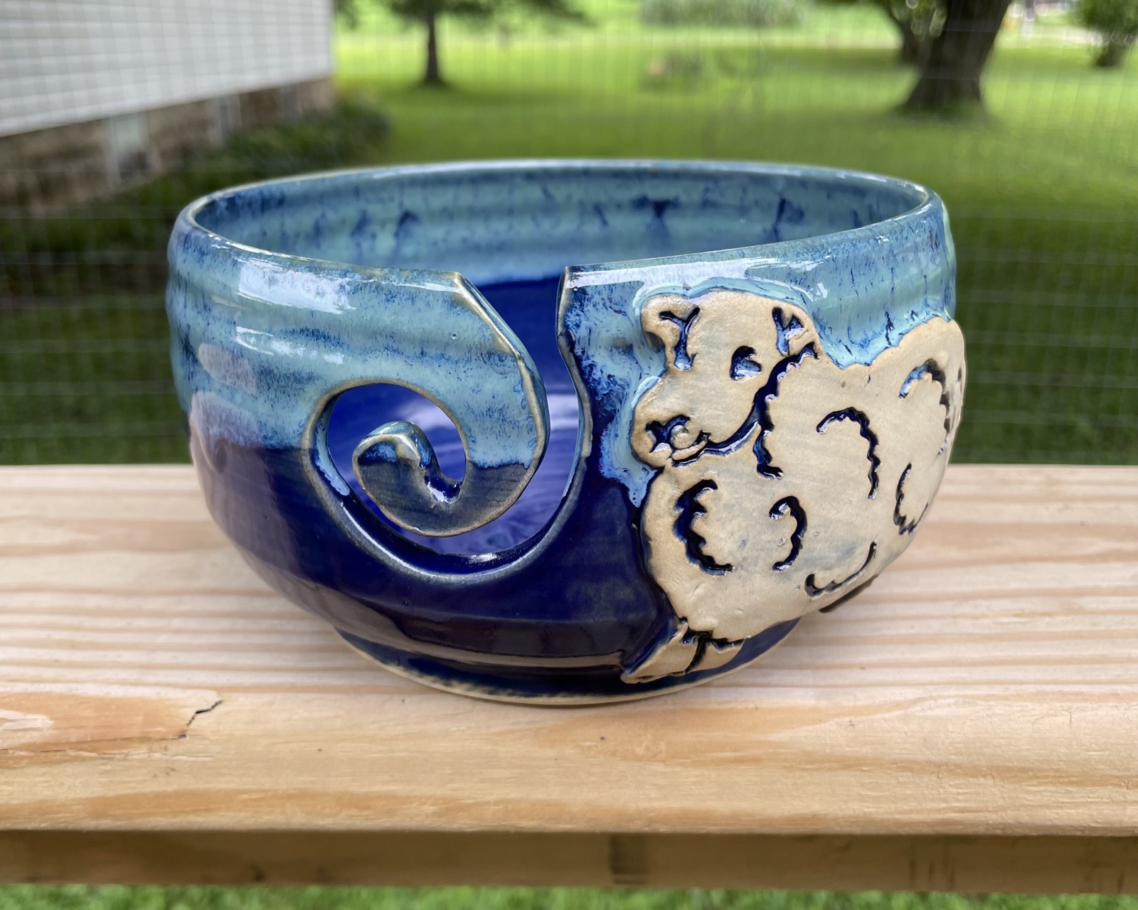 Handmade blue yarn bowl with a sheep applique on a wooden rail.